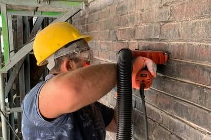 Handwerker arbeitet mit einer Maschine an einer Ziegelwand, trägt Schutzhelm und Schutzbrille.