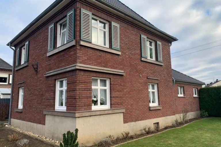 Zweistöckiges, rotbraunes Backsteinhaus mit weißen Fenstern und grünem Garten 